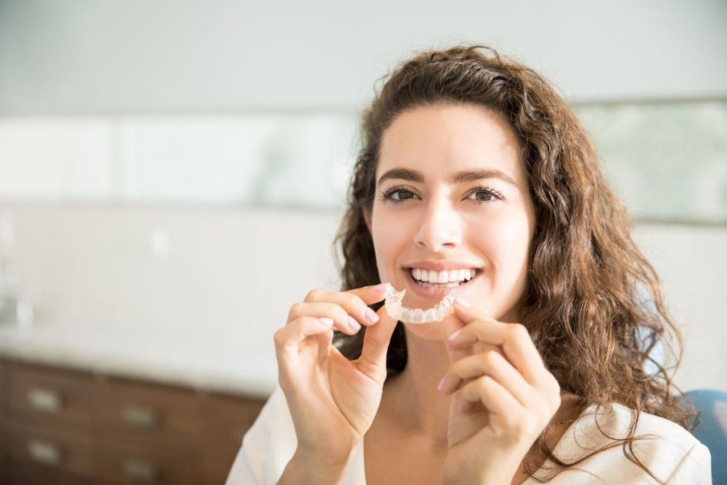 Young woman smiling and holding clear aligners near her mouth Invisible braces & Invisalign Toronto.